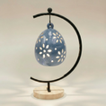 Load image into Gallery viewer, (ice)
Rear view of the ice-blue floral luminary by Ahava Memorials, handcrafted with ashes and shown on its stand with candlelight shining through the ceramic design.